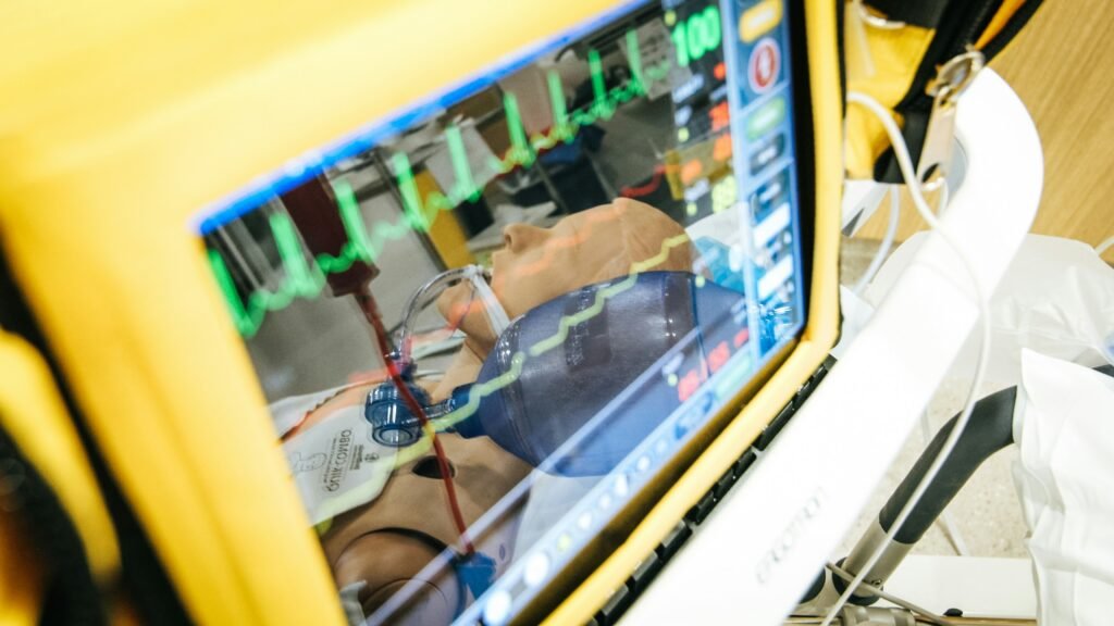 A medical monitor displaying ECG waveforms, reflecting a patient simulation mannequin wearing an oxygen mask in the screen's reflection.